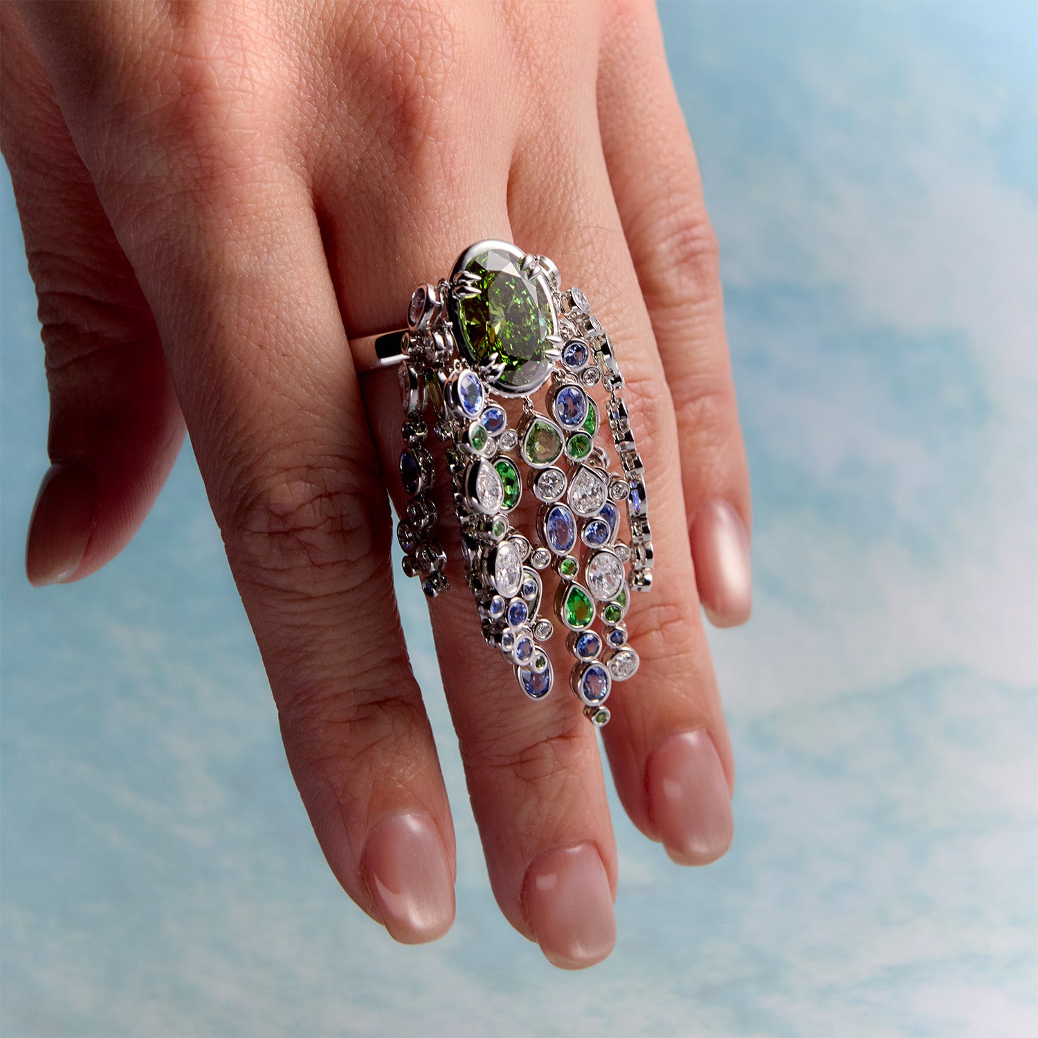 Close-up of a hand wearing a Waterfall ring with streams of blue sapphires, green sapphires, and white diamonds cascade organically across the hand, echoing the rhythm of water catching light as it moves over stone against a blurred background