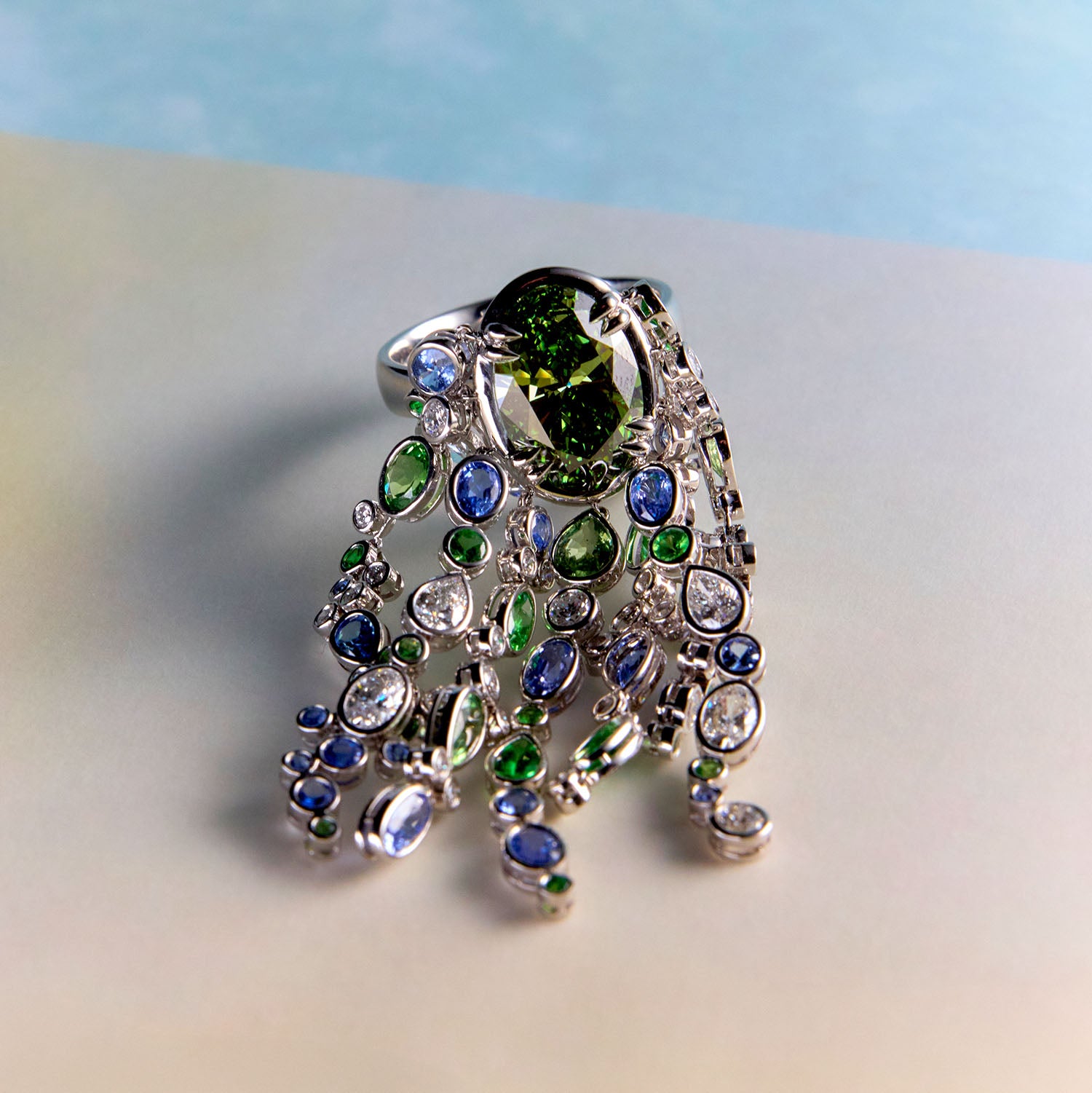 Waterfall ring with streams of blue sapphires, green sapphires, and white diamonds cascade organically across the hand, echoing the rhythm of water catching light as it moves over stone on a light background