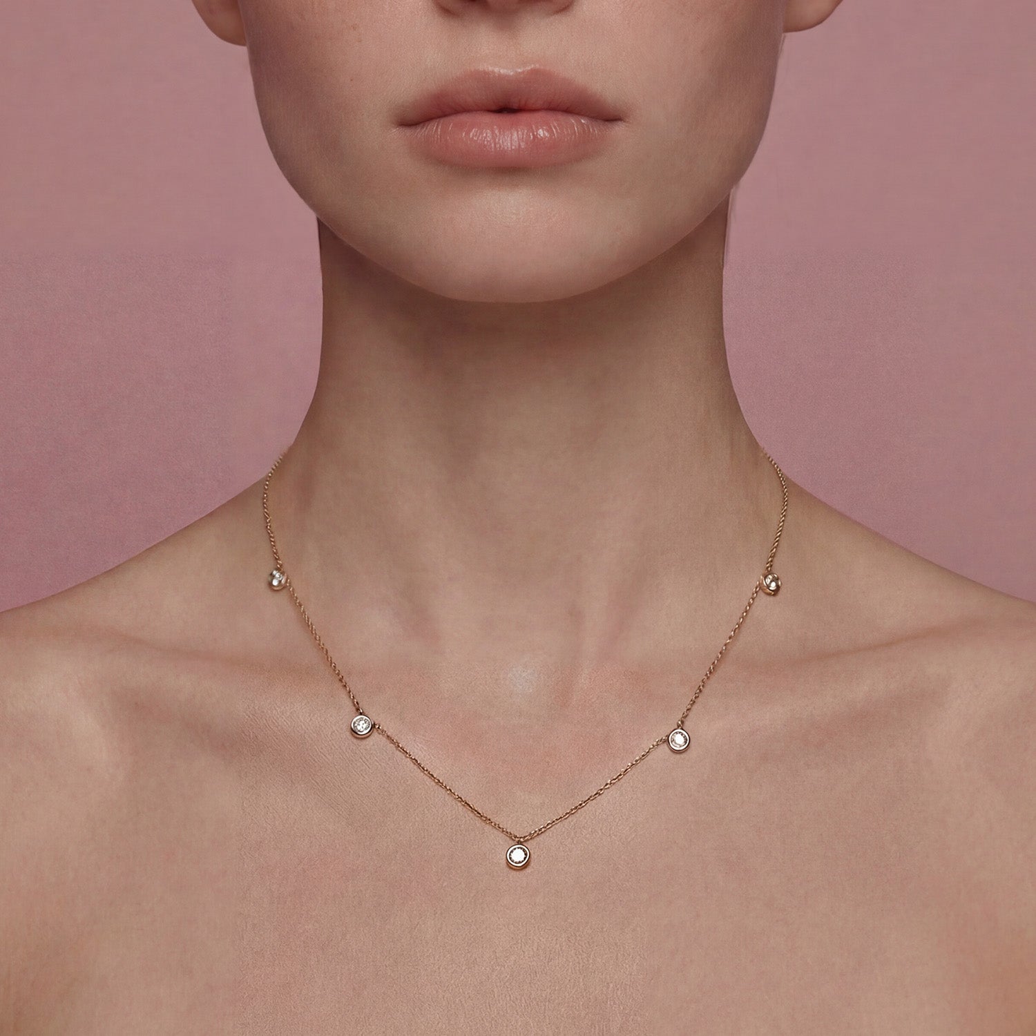 Delicate yellow gold necklace with small round diamond pendants on a person against a pink background